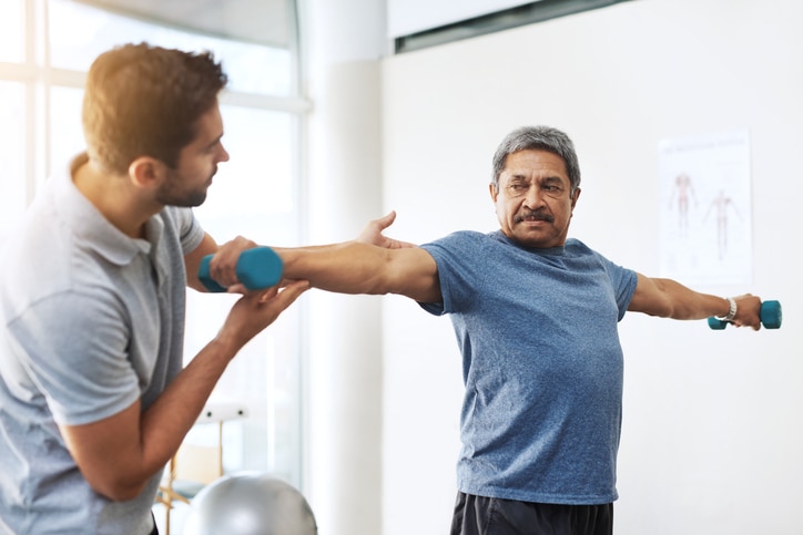 A man holding two dumbbells with outstretched arms, assisted by another man