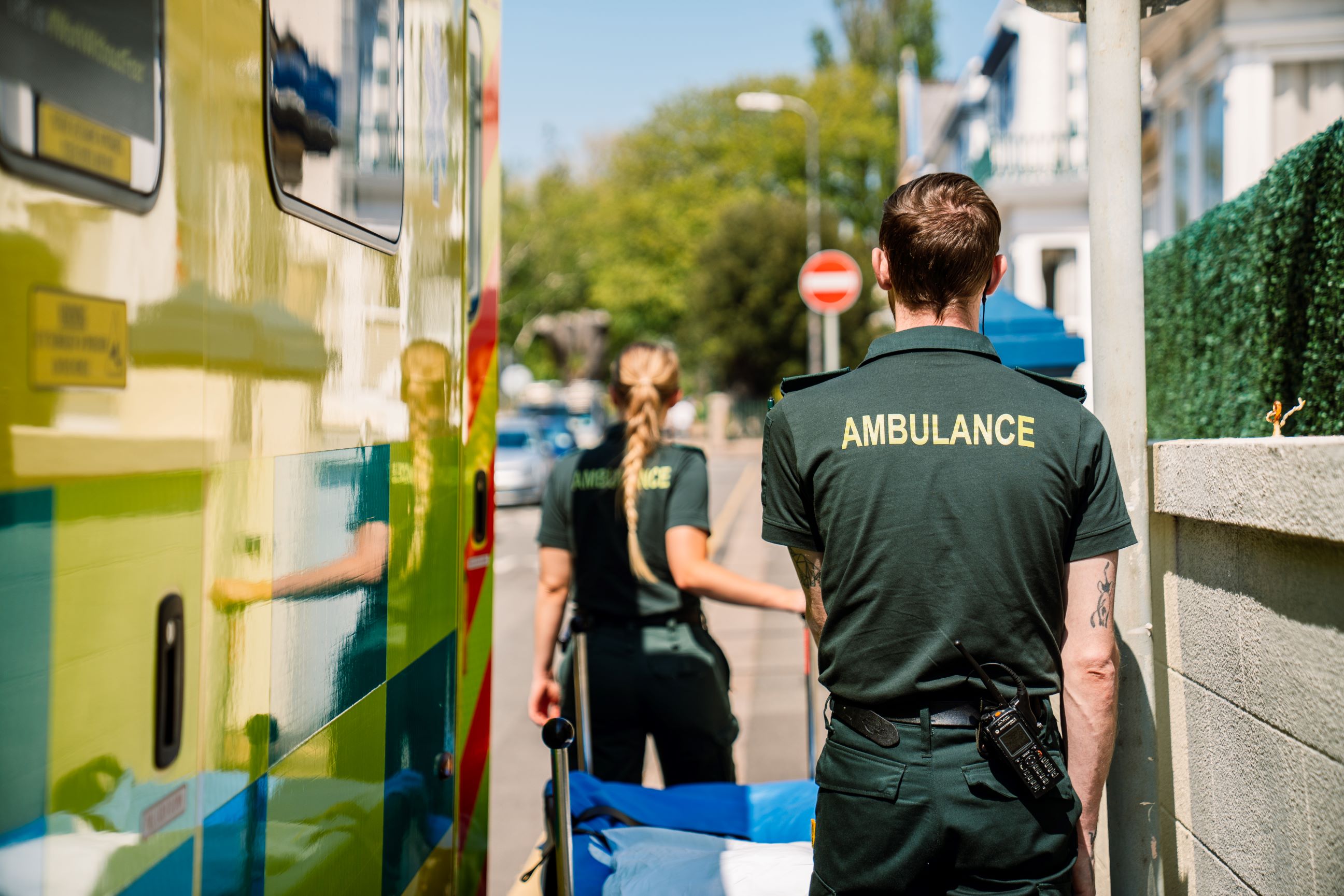 Two paramedics wearing dark green uniforms with 'AMBULANCE' printed on the back, walk along a residential street, pushing a stretcher. An ambulance is parked nearby