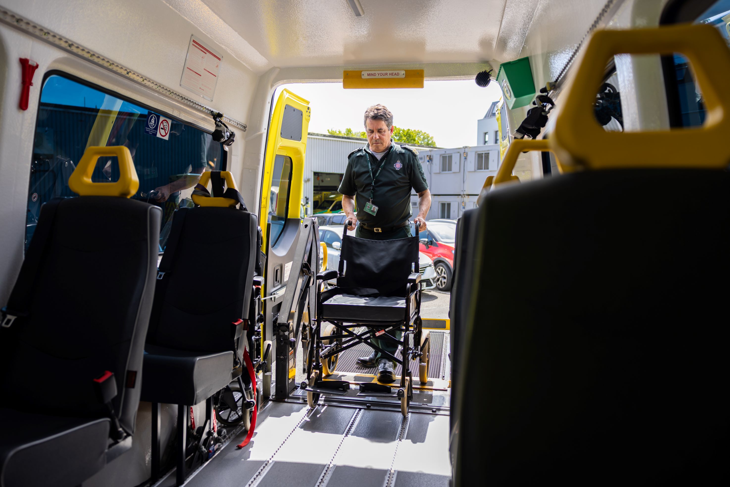 A patient transport employee in dark green uniform stands at the back of a patient transport vehicle, guiding a wheelchair up a ramp into the interior of the vehicle.