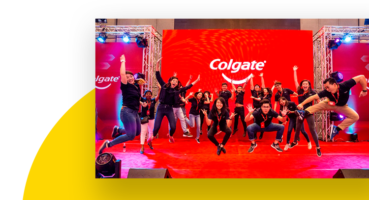 Group of employees jumping and dancing at an annual celebration in front of a red Colgate banner