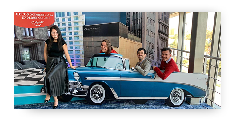 1 employees standing in front of a blue convertible automobile model with 3 other employees sitting in it