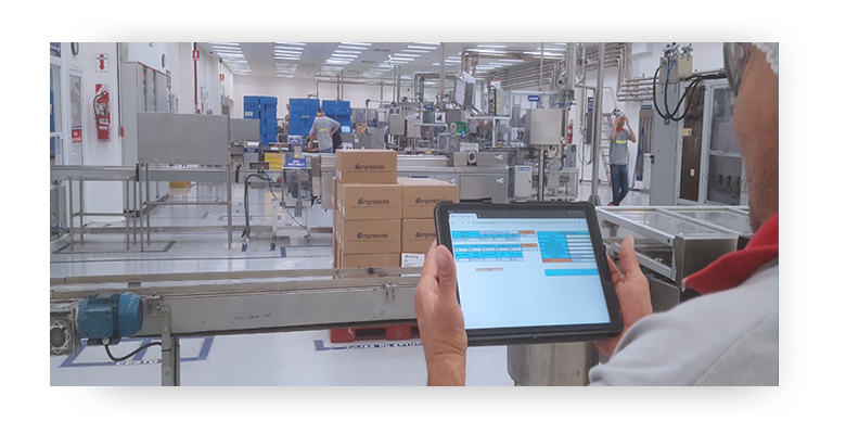 Hands holding an electronic tablet with images on the screen with factory production lines and boxes in the background