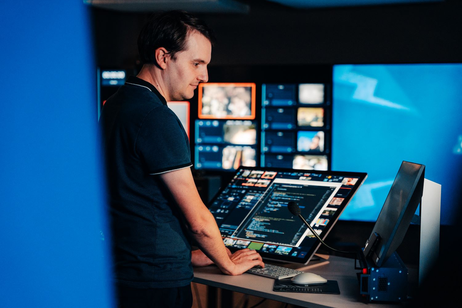 Un homme est debout dans un studio de la RTBF. Les deux mains posées sur le clavier, il regarde un des deux écrans de travail devant lui. Plusieurs autres écrans lumineux sont visibles en arrière-plan.