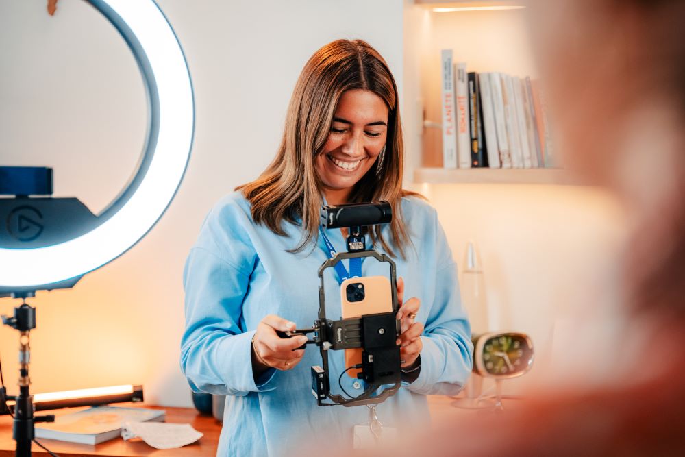 Dans un studio de la RTBF, une femme, souriante, est debout et manipule une steadicam pour smartphone. Un Ring Light est visible en arrière-plan.