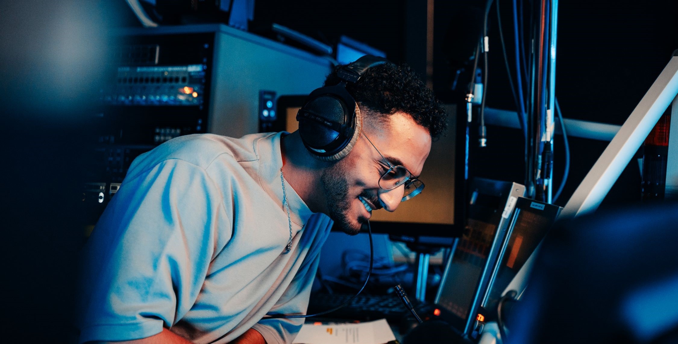 Dans un studio de la RTBF, un homme souriant, un casque audio sur les oreilles, est assis devant deux écrans d'ordinateur. Un panneau de réglages est visible en arrière-plan.