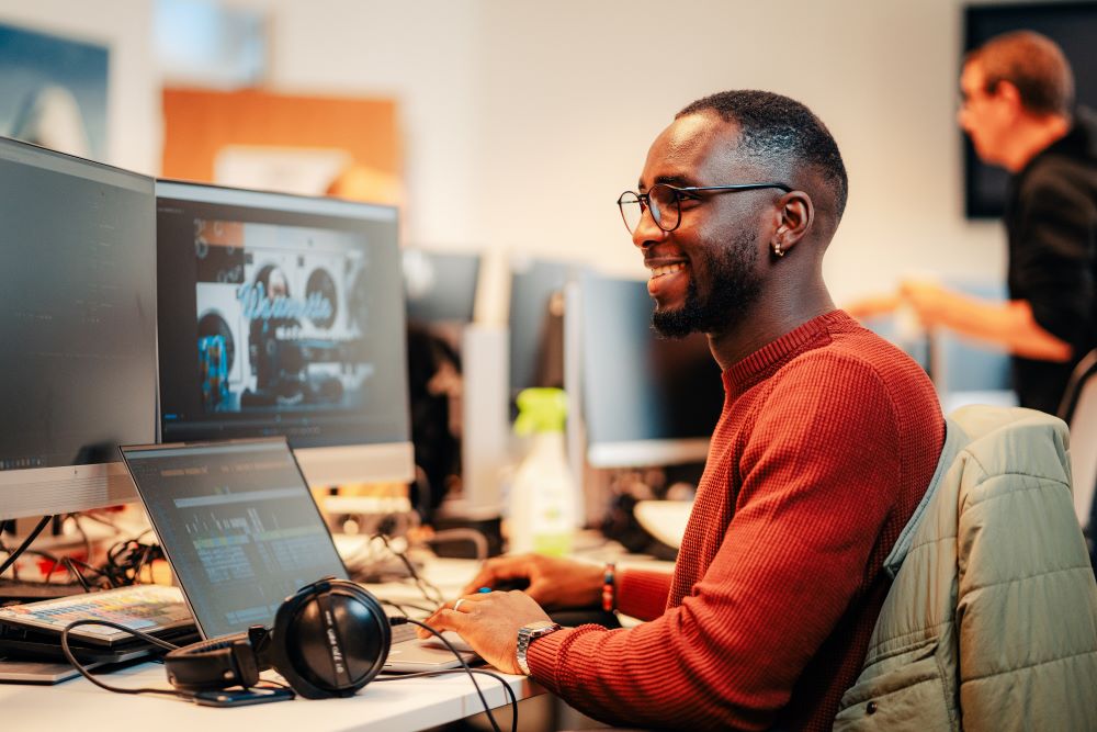 Dans un espace de travail, un homme est assis face à trois écrans d'ordinateur. Il tape sur un clavier en souriant. Plusieurs ordinateurs et un homme, debout, sont visibles en arrière-plan.