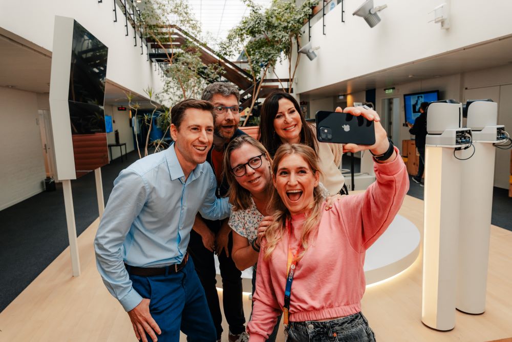 Dans les locaux de la RTBF, un groupe souriant de deux hommes et trois femmes, se prend en photo avec un smartphone. 