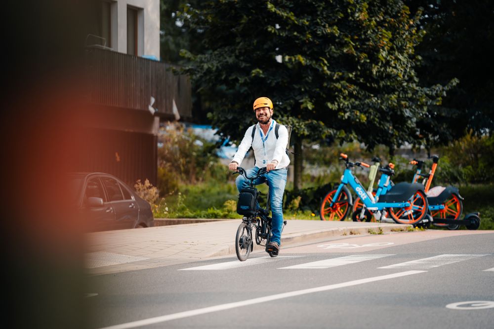 A l'extérieur, dans un environnement verdoyant, à proximité d'une habitation, un homme portant un casque et un sac à dos roule à vélo sur une piste cyclable.