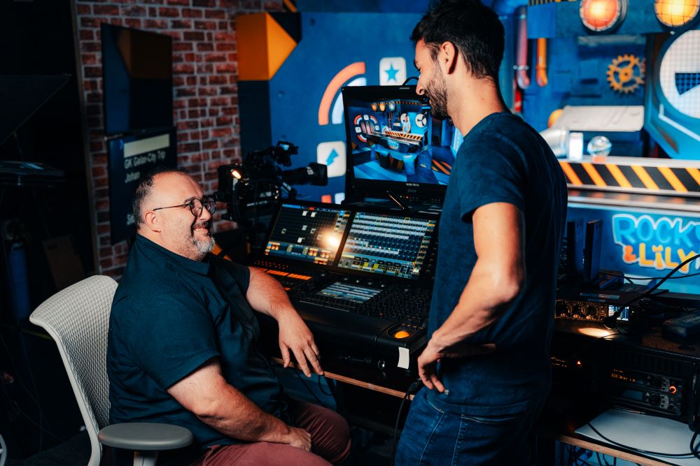 Deux hommes, un assis et un autre debout, discutent dans un studio de la RTBF. Plusieurs écrans et appareils de réglages se trouvent devant eux.