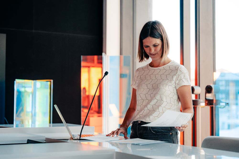 Dans un studio, une femme, debout, regarde plusieurs documents déposés sur un bureau (plateau de télévision). Un micro et un ordinateur portable sont posés devant elle.