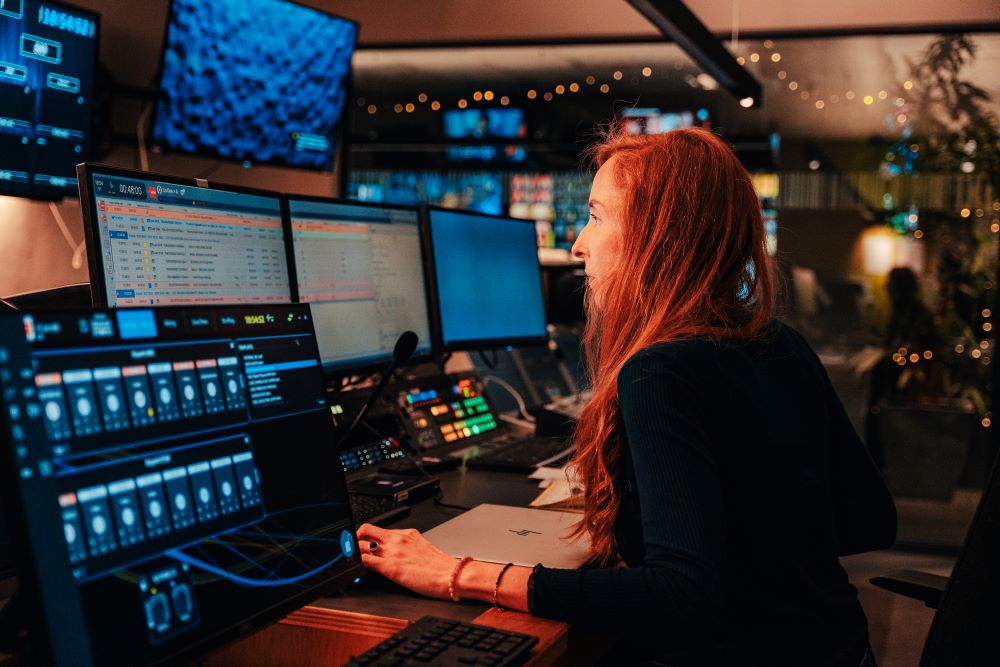 Une femme, de profil, travaille dans un studio de la RTBF. Plusieurs écrans, clavier et boutons de réglages sont devant elle. Des écrans sont visibles en arrière-plan.