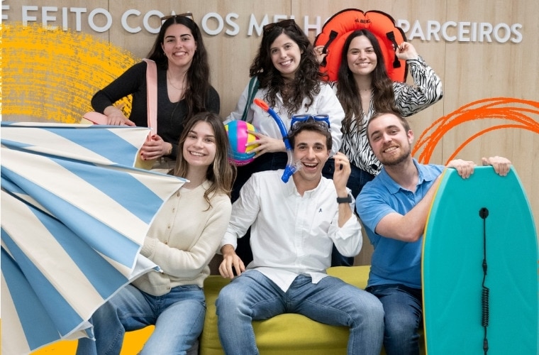 Um grupo de seis pessoas posa com vários itens relacionados à praia. O fundo tem um texto que diz "FEITO COM OS MELHORES PARCEIROS." Os itens incluem um grande guarda-sol listrado azul e branco, uma bola de praia colorida, uma boia insuflável e uma prancha de bodyboard. A imagem promove os estágios de verão Call for Summer da MC Sonae.