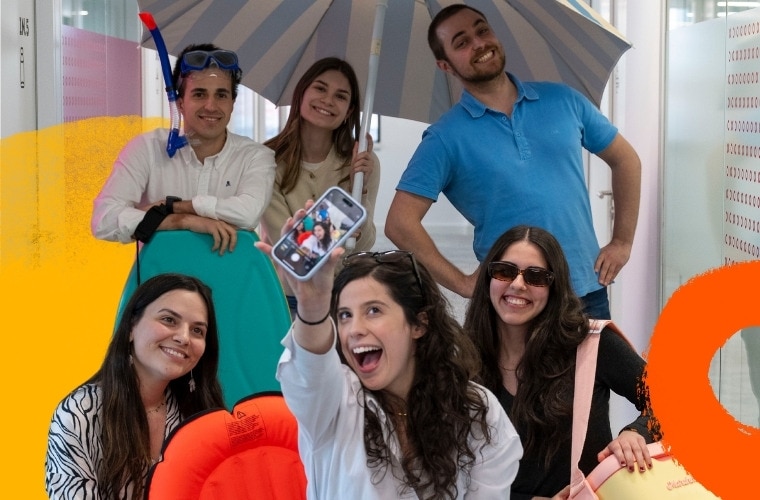 Um grupo de seis pessoas posa juntas. Uma pessoa na frente está segurando um smartphone, exibindo uma foto no ecrã.  O grupo parece estar em um ambiente interno, e alguns indivíduos estão segurando itens relacionados à praia, como um guarda-sol, equipamentos de mergulho e dispositivos insufláveis de flutuação. Imagem promove o programa de estágios de verão Call For Summer da MC Sonae.