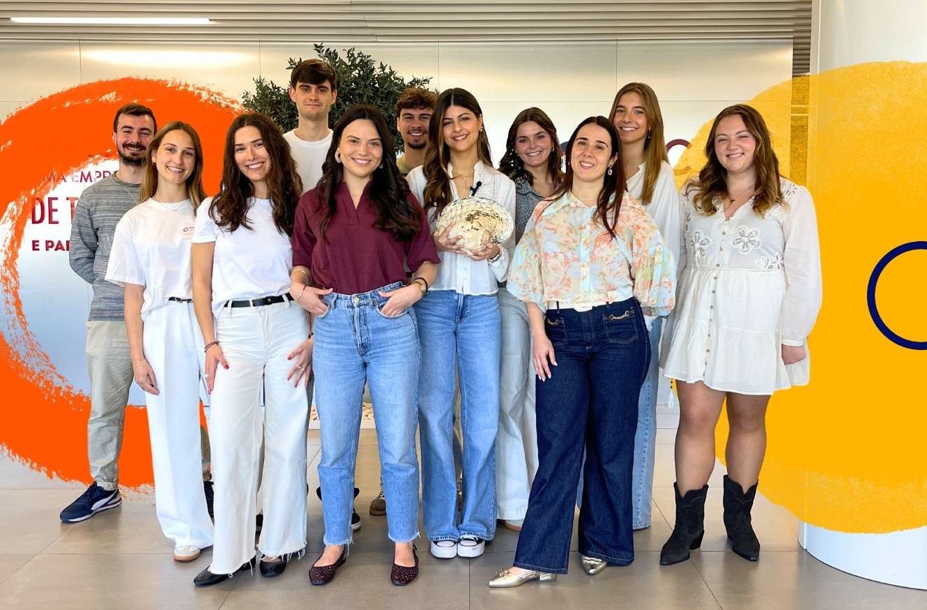 Fotografia de grupo com várias pessoas de pé, lado a lado, num espaço interior corporativo. O grupo está posicionado à frente de uma parede clara com elementos gráficos decorativos em tons de laranja e amarelo. Uma das pessoas segura uma grande concha natural ao centro da imagem.