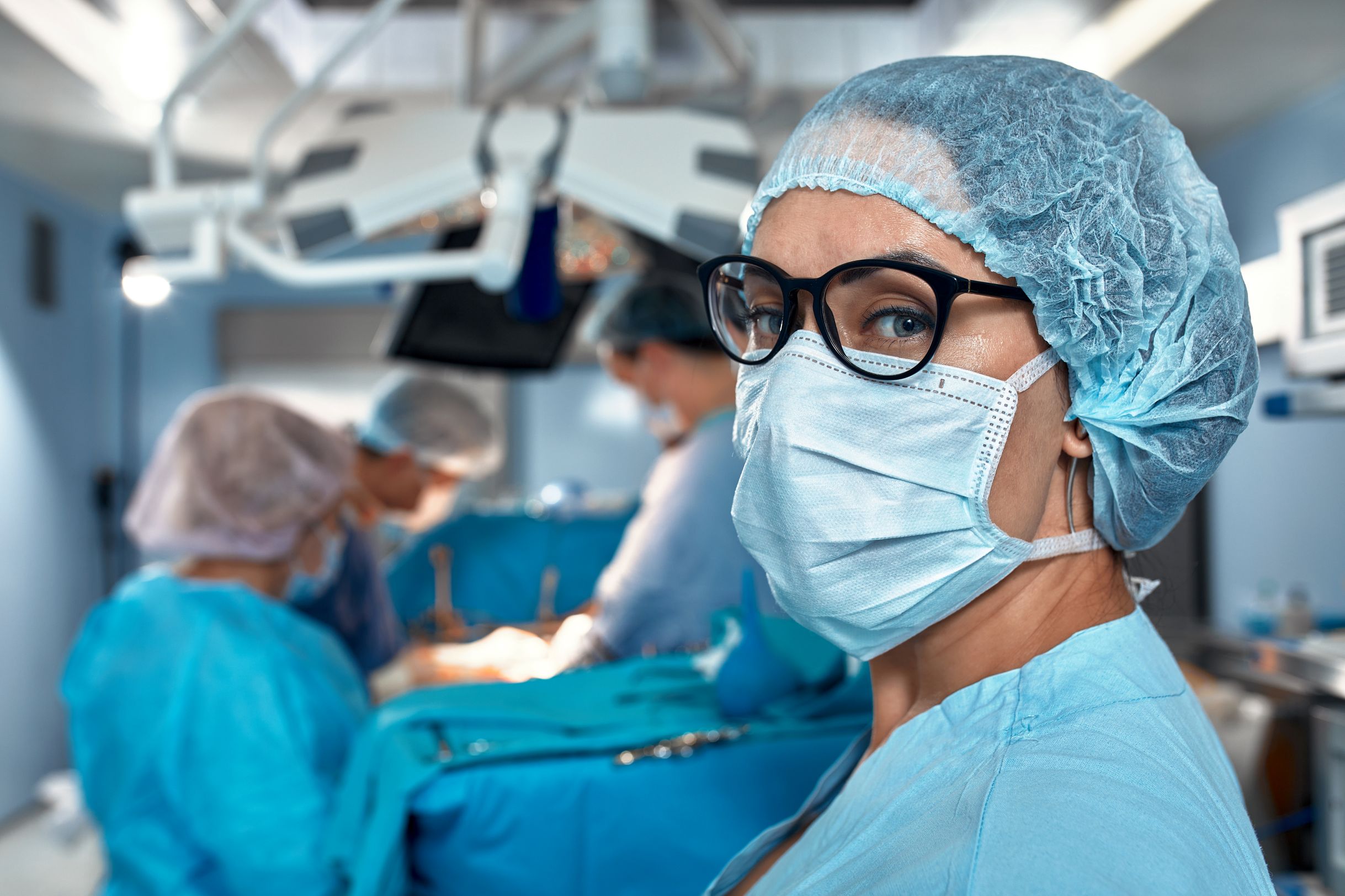 Healthcare professional in surgical attire standing in an operating theatre while a surgical team works in the background.