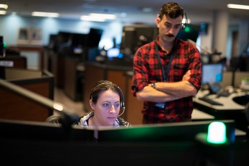 Une répartitrice assise à son poste de travail. Un répartiteur masculin se tient à côté de la femme et regarde son écran d'ordinateur.