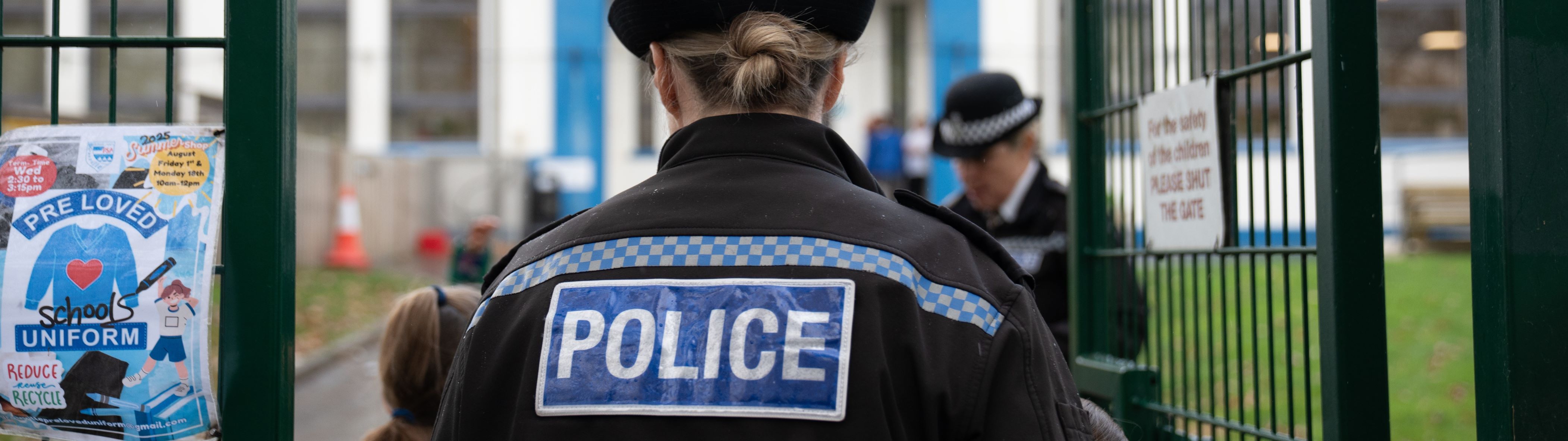 Two female police officers in black uniforms with 'POLICE' written on the back walk through a gate into a school. They are wearing traditional British-style police hats with a checkered band.