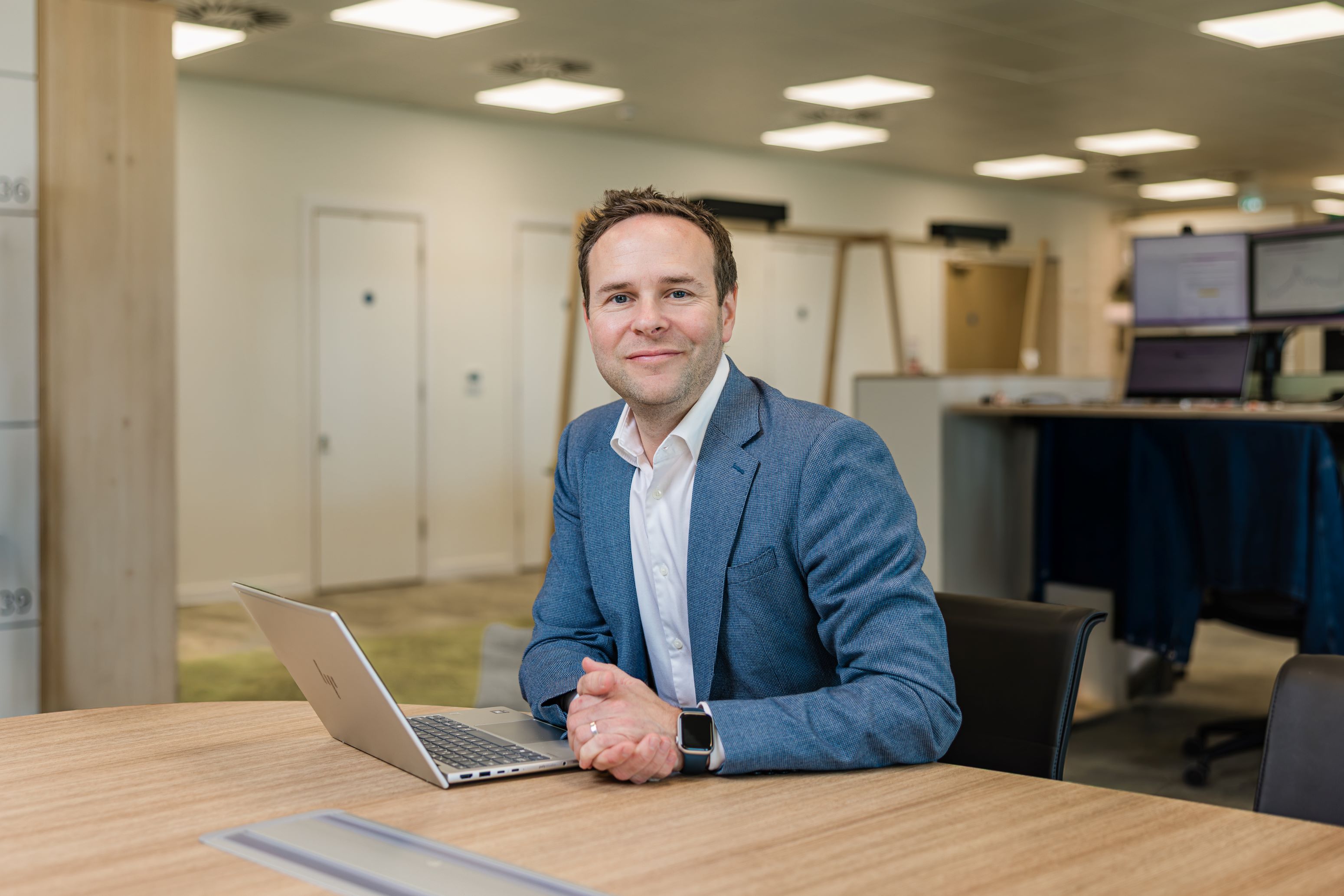A man with short, light brown hair, and a smile is wearing a blue suit jacket with a white shirt. It is Paul Wylie, CEO of Government of Jersey. He is sitting at a desk in the Government of Jersey office.