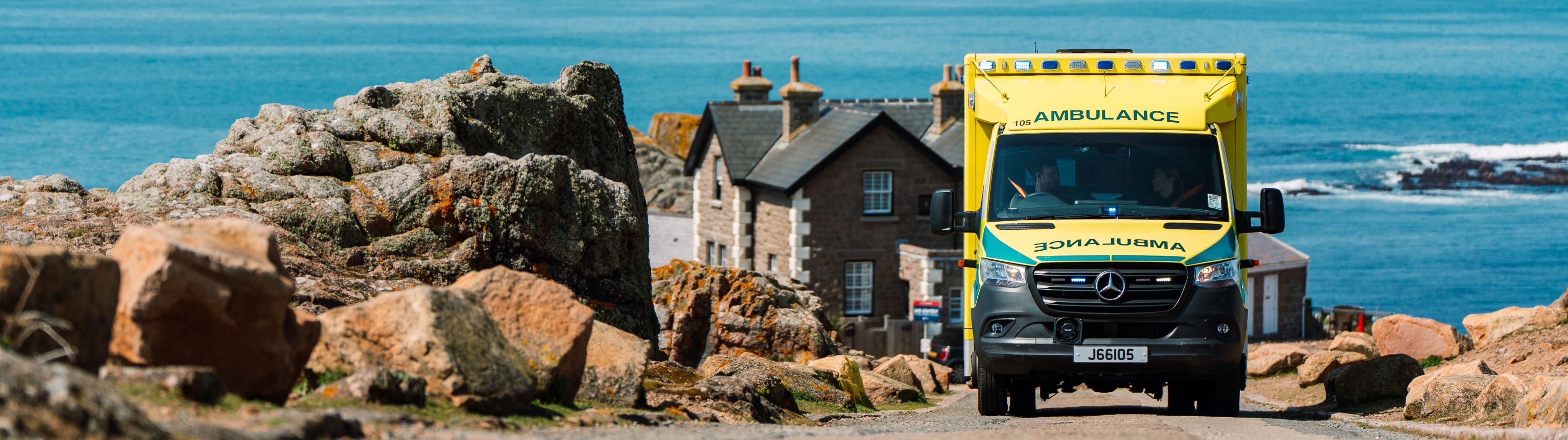 A yellow ambulance drives along a beautiful coastal road in St Ouen (Jersey), flanked by rocky terrain and a traditional granite building. The sea and a bright blue sky stretches out in the background.