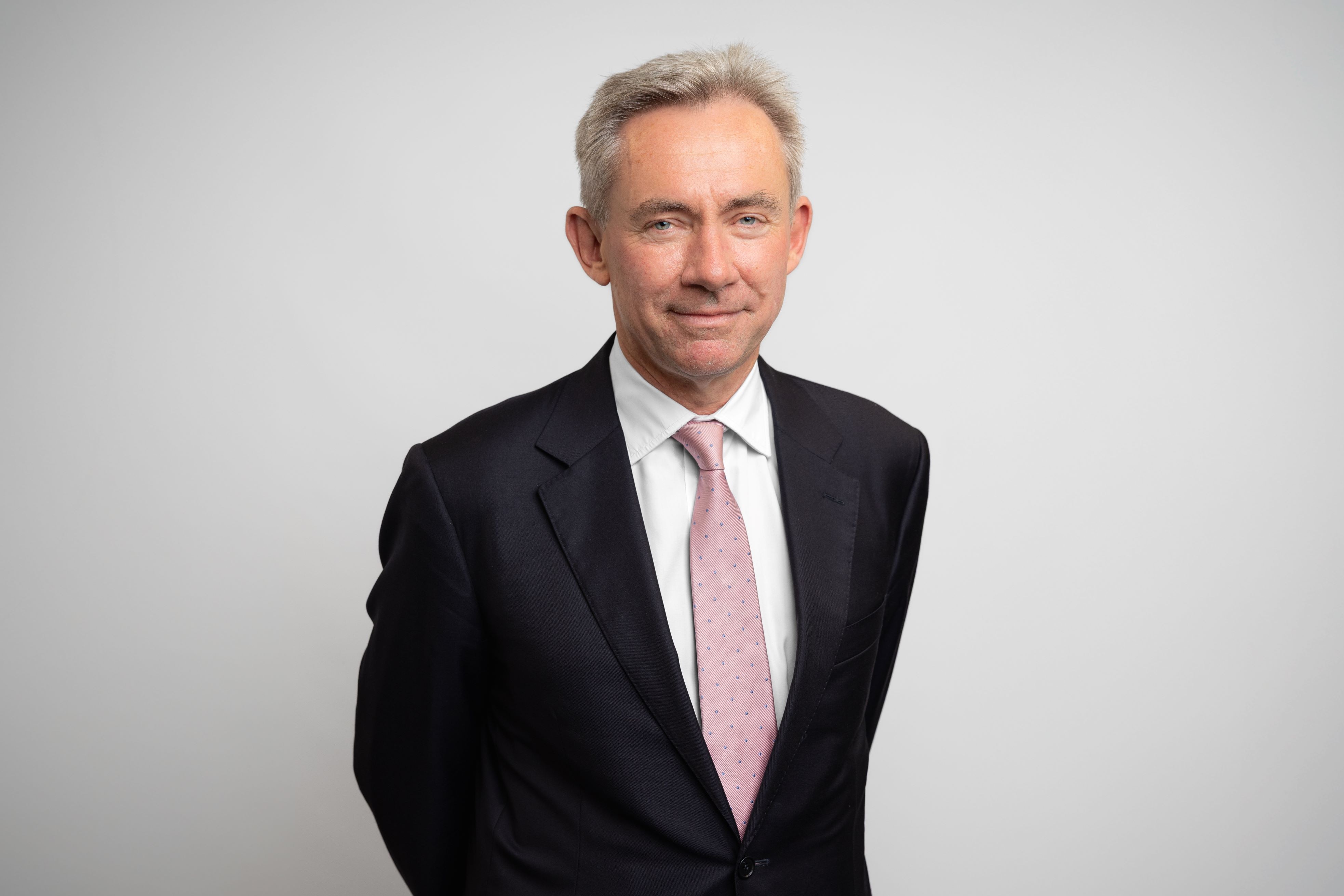 A professional head shot of a man in a black suit, white shirt, and pink tie, standing against a light grey background.