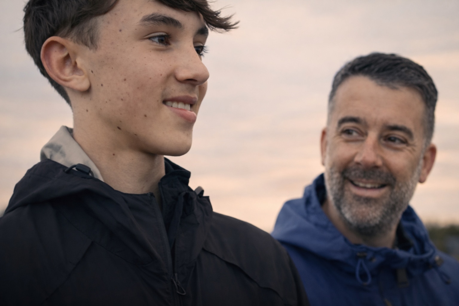 A man in a blue jumper is looking at a boy on the left side of the image. they are outdoors, smiling.