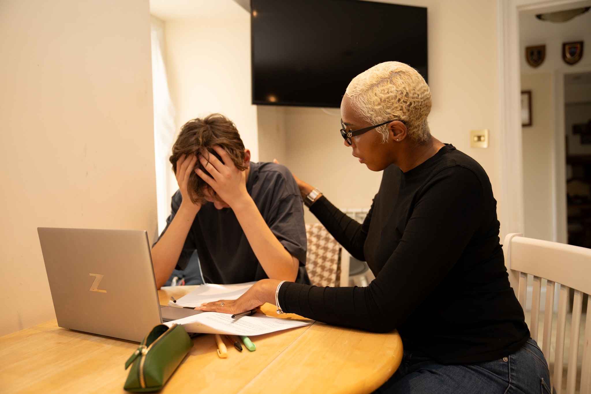 An adult sitting beside a young person at a table with a laptop and paperwork.