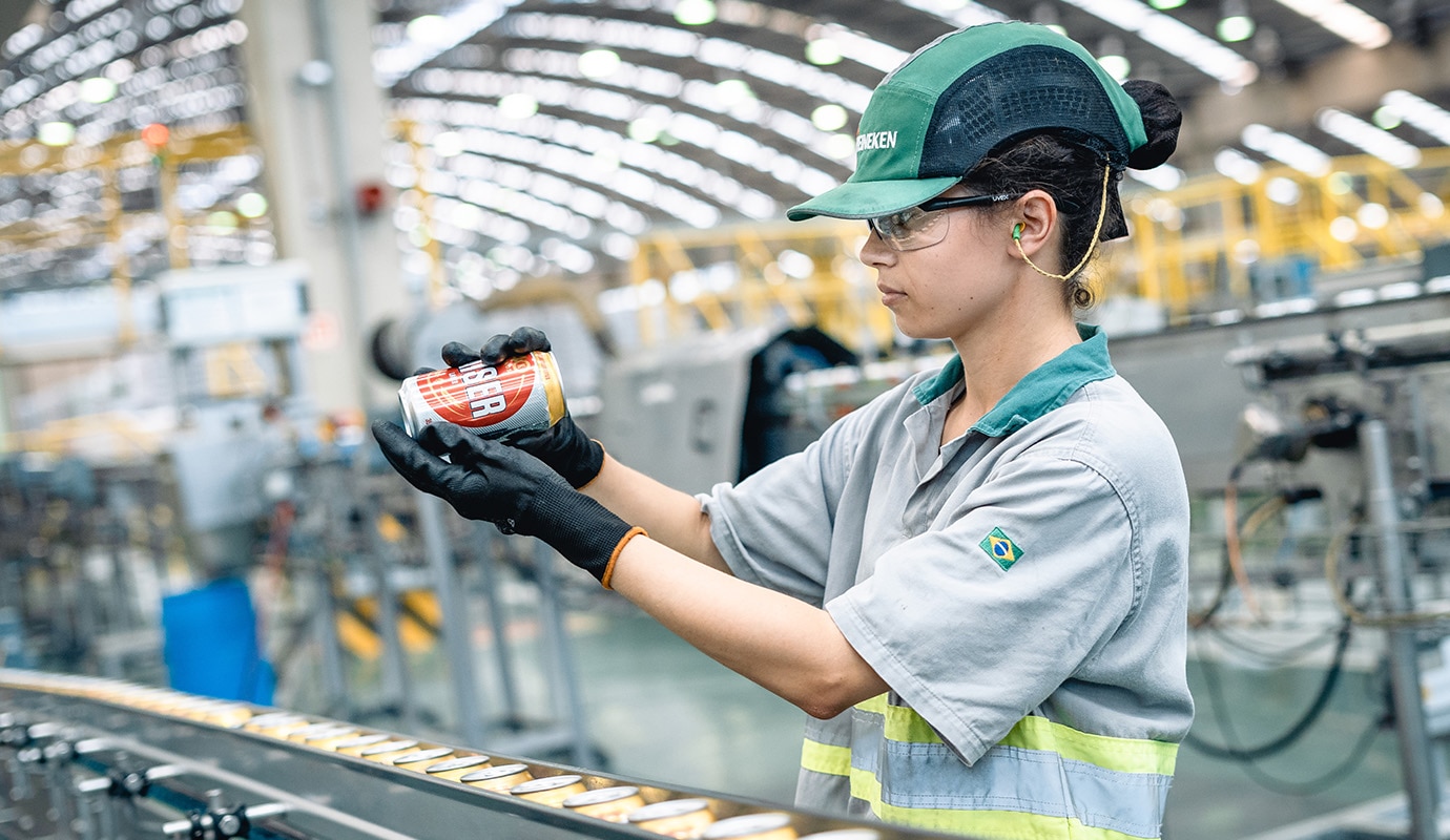 Person working in a factory wearing a HEINEKEN cap and inspecting a drinks can.