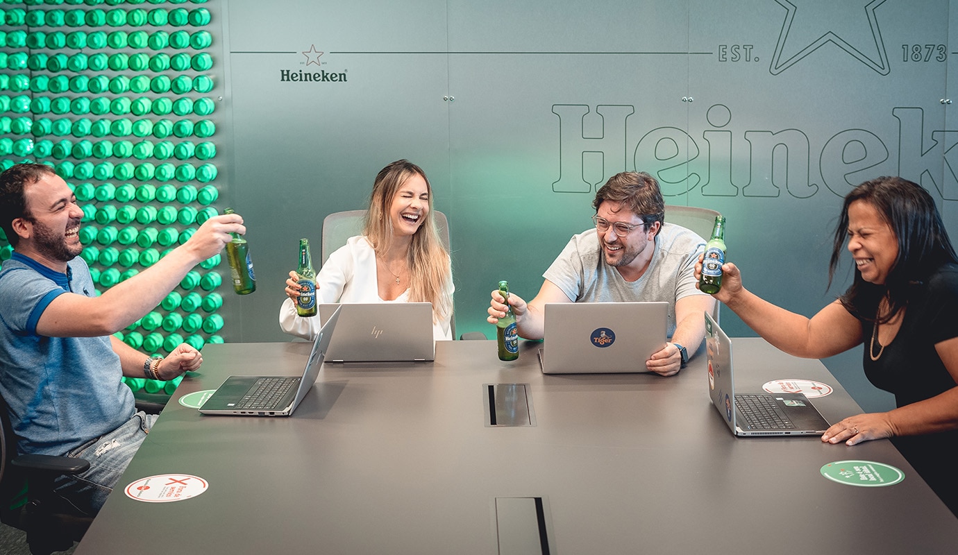 Four HEINEKEN colleagues in a meeting room, sat at a table holding bottles of Heineken 0.0 and smiling.