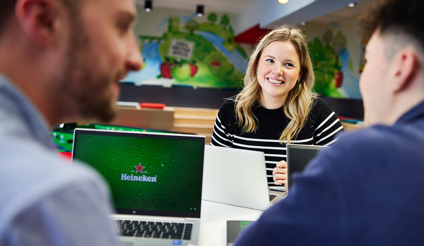 Three young HEINEKEN Digital Graduate colleagues sat smiling and speaking to each other in a HEINEKEN office next to a laptop.