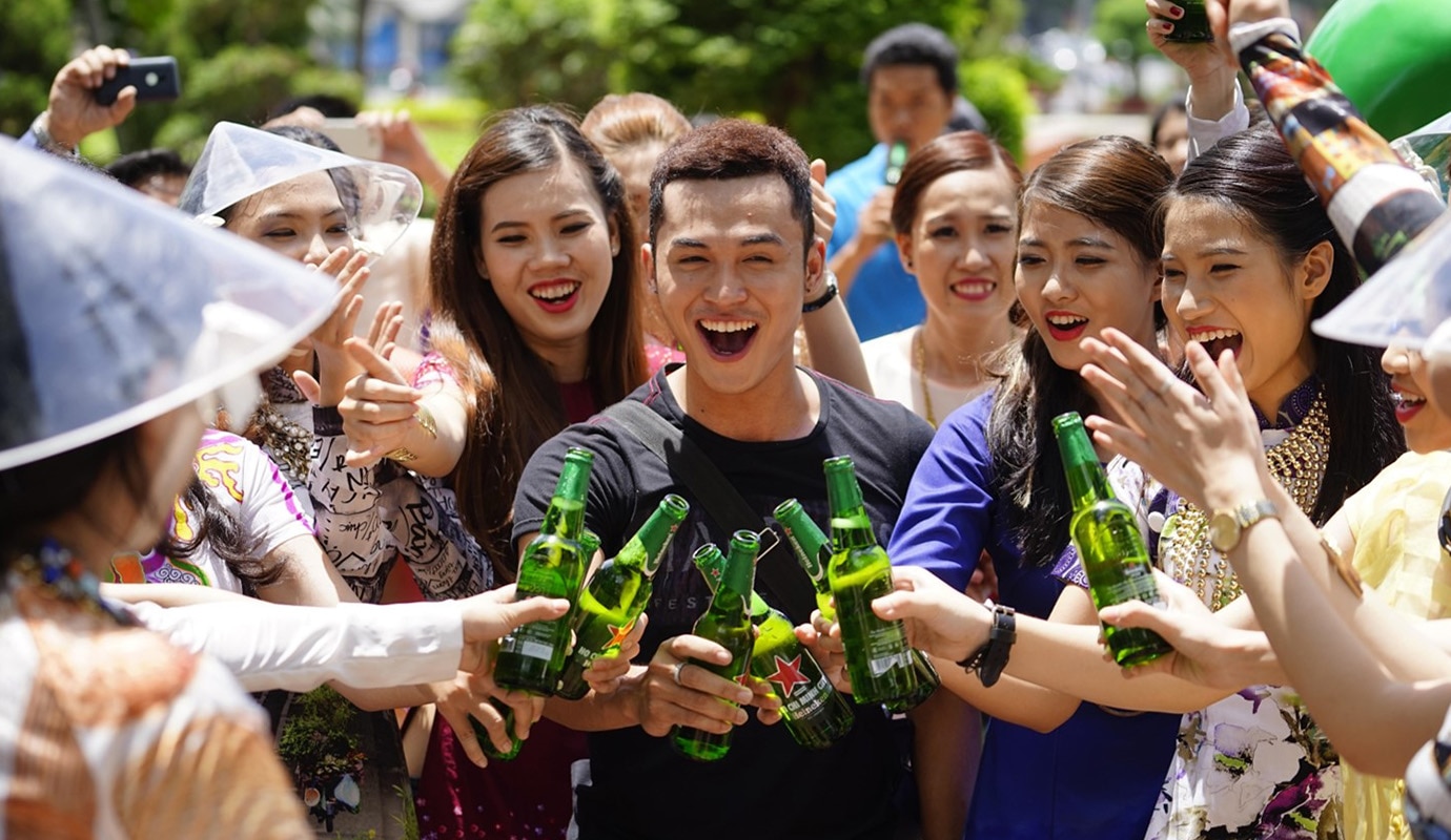 Group of people outdoors in bright weather, all holding bottles of Heineken beer and 'cheersing' together.