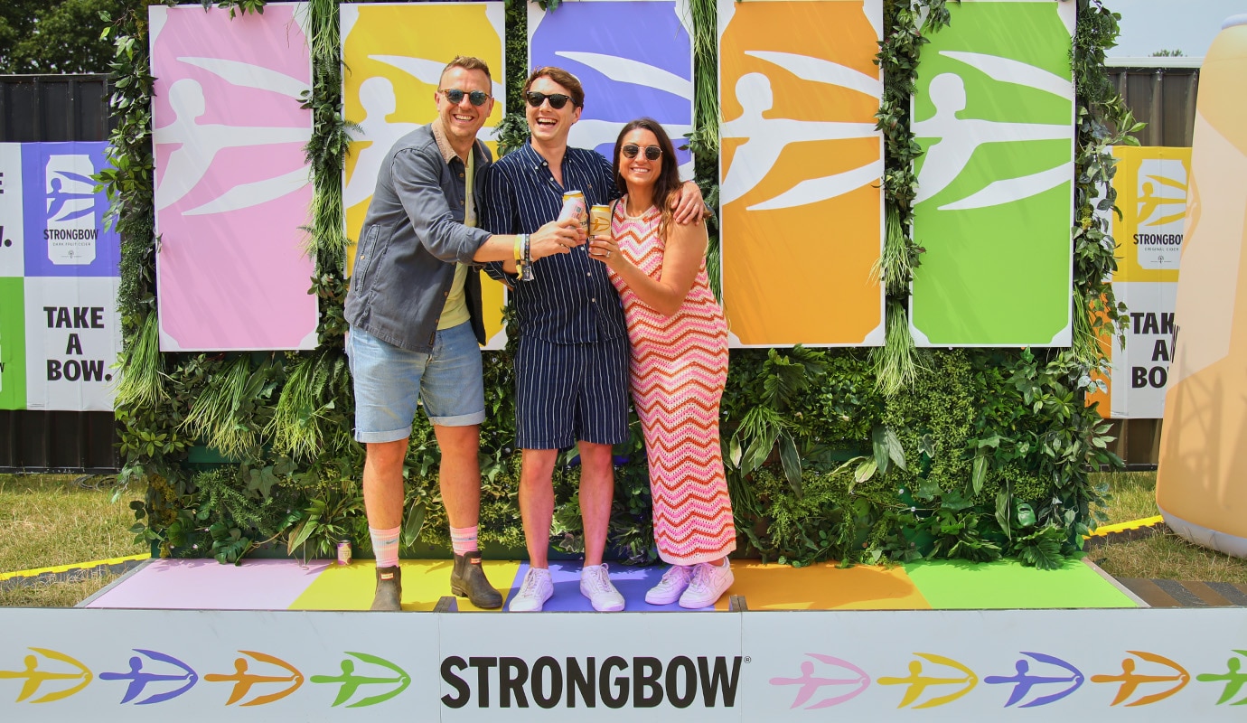 Three colleagues stood in front of a branded Strongbow backdrop at the Isle of Wight festival, smiling.