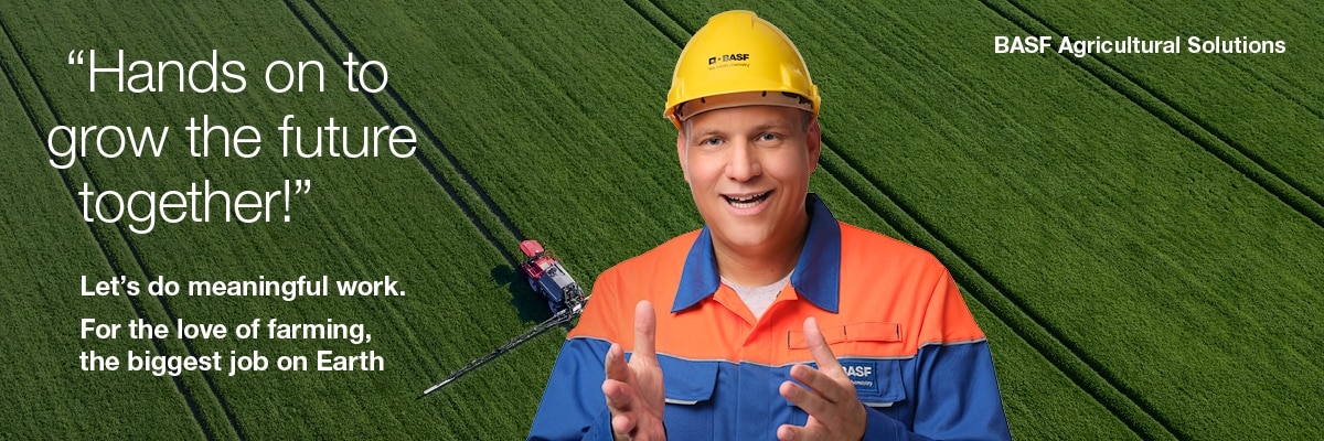 A person wearing a yellow safety helmet and orange-blue work clothes with the BASF logo stands in front of a green field with visible rows of crops. Agricultural machinery can be seen in the background. On the left, the English text reads: Hands on to grow the future together!‘, ‚Let's do meaningful work. For the love of farming, the biggest job on Earth‘ and ‚BASF Agricultural Solutions