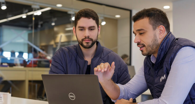 Dois homens brancos conversam enquanto observam a tela do computador em uma reunião dentro de um escritório. A sala é bem iluminada e tem uma parede de vidro ao fundo que da vista ao restante da empresa.