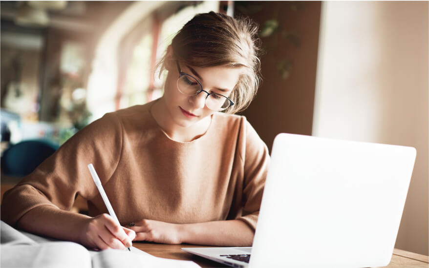 Uma mulher branca loira de óculos e uma blusa
          marrom, com a cabeça abaixada enquanto olha para o papel no qual está escrevendo e com um notebook em sua frente
          na mesa.