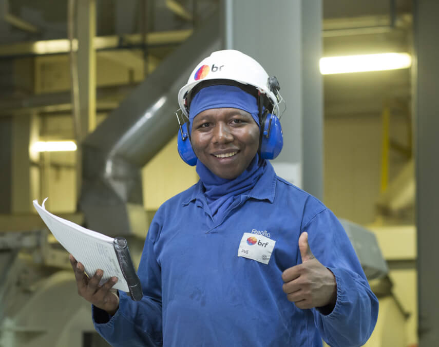 Homem negro sorridente vestindo um uniforme azul da BRF, utilizando
        equipamentos de EPI. Segurando um caderno e fazendo um gesto de afirmativo com o polegar
