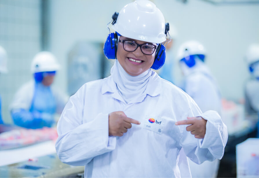 Mulher negra sorridente, aponta para o logotipo da BRF em sua roupa de trabalho. Ela está usando equipamentos de proteção individual (EPI), como óculos de proteção, protetor auditivo,  capacete e jaleco. A imagem foi tirada dentro de uma fábrica de alimentos.