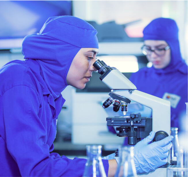 Uma mulher branca vestida com uniforme e luvas azuis, olhando através de um microscópio com frascos químicos à frente. Ao fundo, uma mulher branca com óculos de grau, também de uniforme azul.