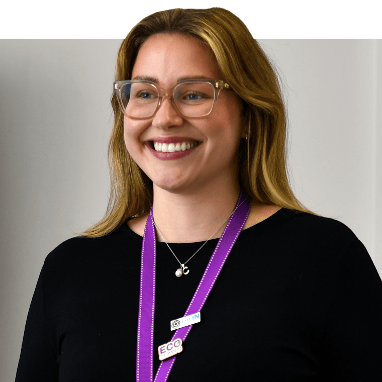 A colleague with light coloured hair and clear frames glasses, wearing a purple lanyard and black top smiling while looking away from the camera. 