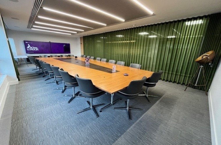 A meeting room with a large table in the centre and a green coloured wall with TV screens on the far wall displaying the Royal London logo.