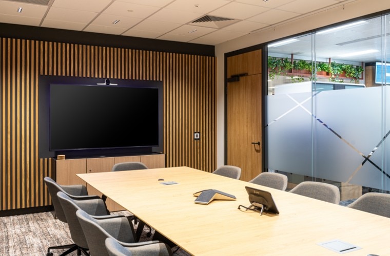 A meeting room with a large TV on the wall and a table in the centre surrounded by chairs with a floor to ceiling glass wall.