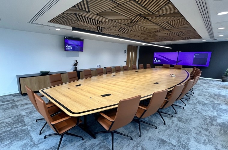 A meeting room with a large table in the centre with a wooden panel above, with TV screens on the far wall displaying the Royal London logo.