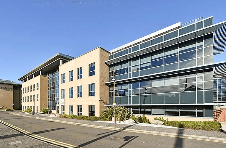 A modern looking stone and glass building with green bushes and shrubs outside.
