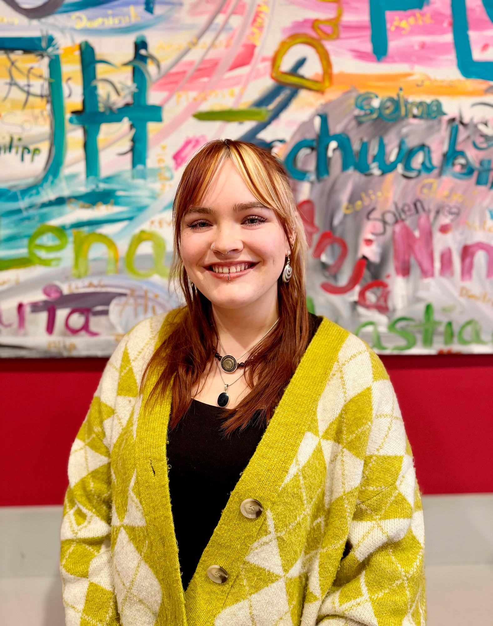 Eine Praktikantin steht vor einer bunten Wand. Sie blickt und lächelt direkt in die Kamera.