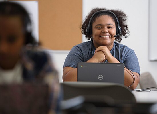 Young student sits at desk with open laptop wearing headset and smiling