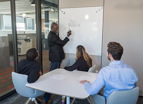 Black man stands at a white board and draws a pie chart while others pay attention