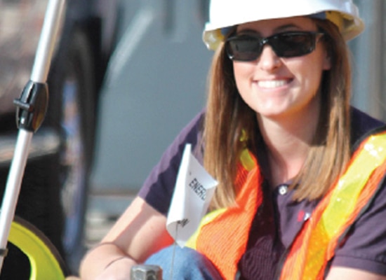 Woman in Xcel Energy hard hat, safety vest, and sunglasses smiles at camera