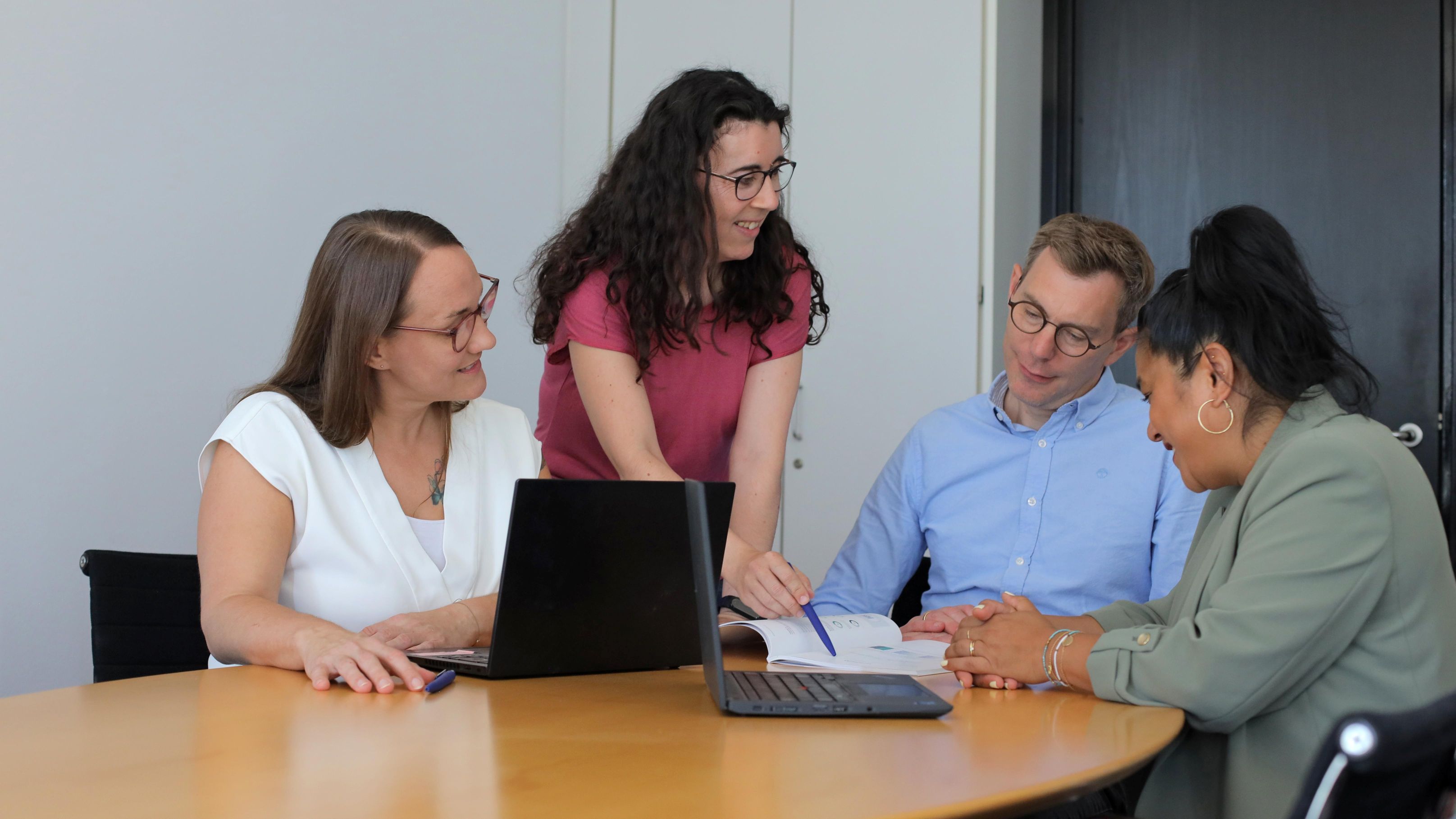 Zwei Kolleginnen und ein Kollege sitzen an einem Besprechungstisch. Eine Kollegin steht bei ihnen. Sie besprechen sich gut gelaunt.