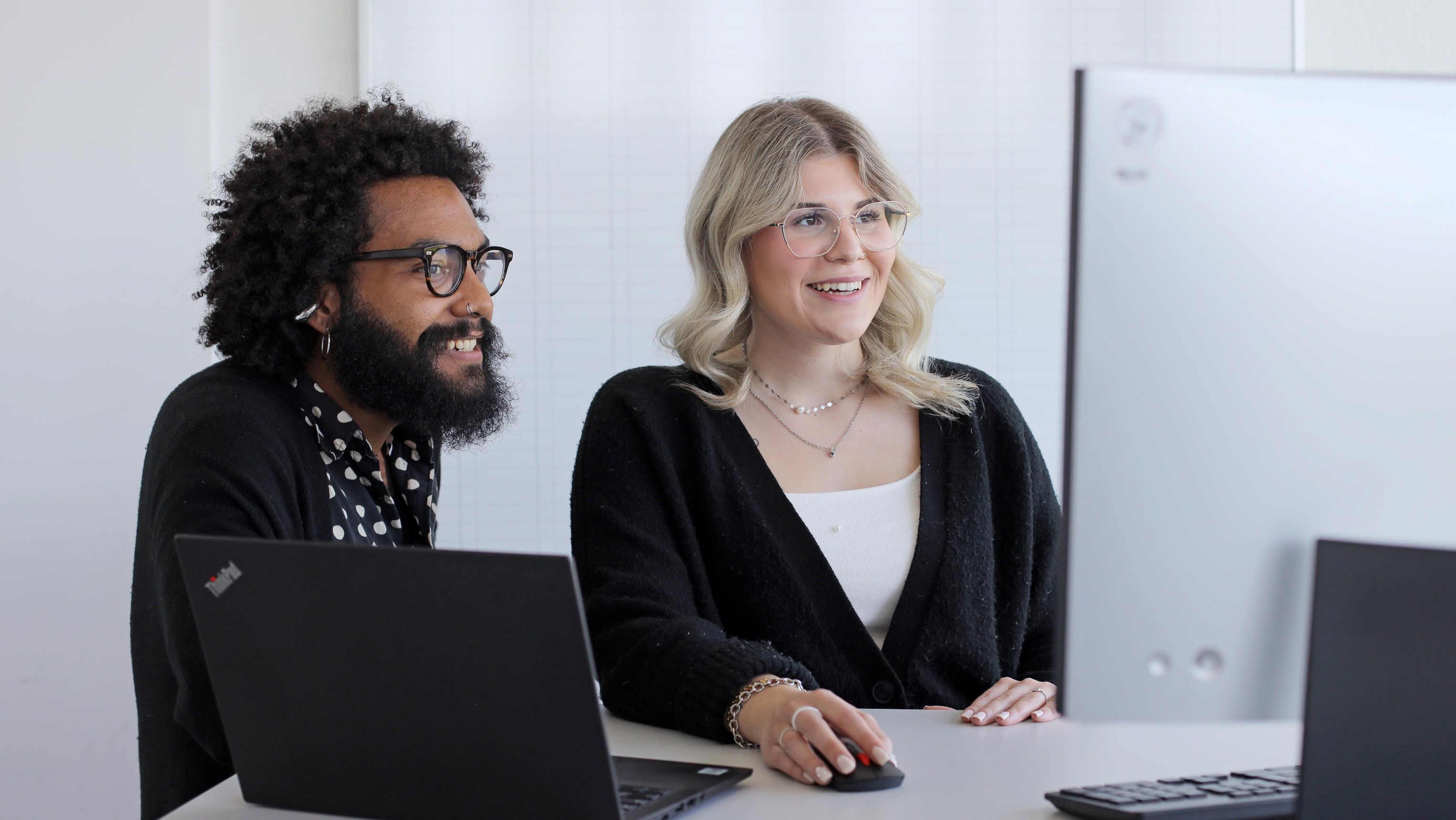Ein Auszubildender und eine Auszubildende stehen gut gelaunt am Schreibtisch und schauen gemeinsam auf einen Bildschirm.