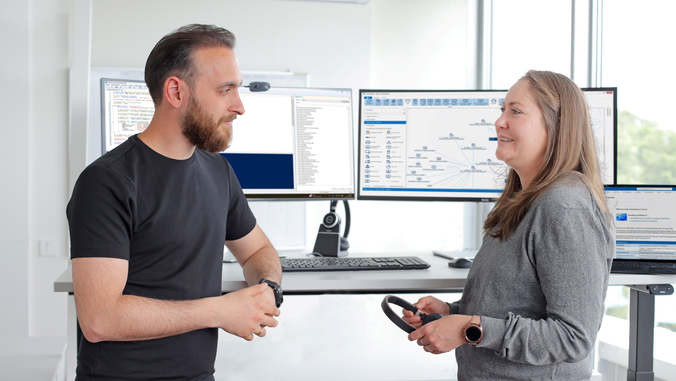 Ein Kollege und eine Kollegin stehen am Schreibtisch und untehalten sich gut gelaunt. Auf dem Schreibtisch stehen 2 große gebogene Bildschirme.