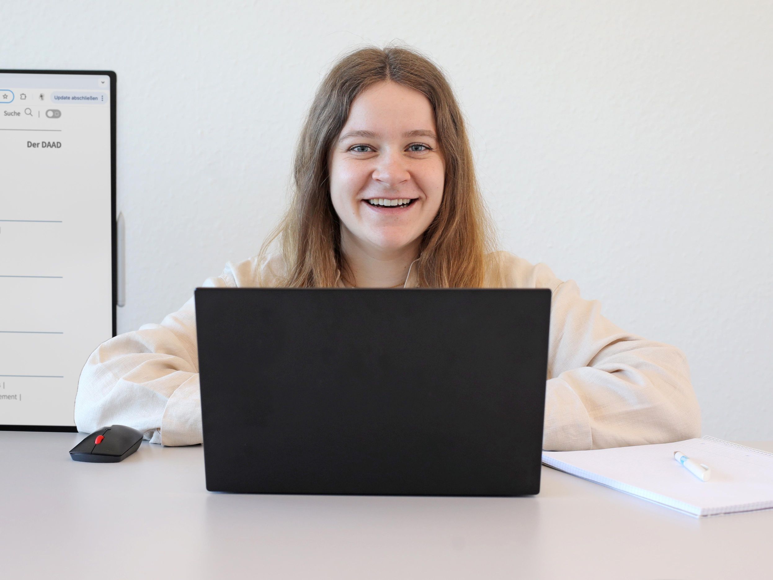 Eine Kollegin sitzt an einem Schreibtisch und lächelt frontal in die Kamera. Hinter ihr ist ein großer Surface-Bildschirm zu sehen.